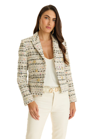 A woman in the Gabrielle Double Breasted Tweed Blazer - Ecru Multi over a white top and cream pants stands with one hand by her side, wearing gold earrings, a belt, and displaying long dark hair.