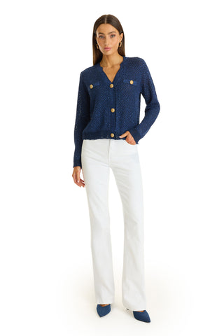 A woman poses against a white background in the Shannon Sequin Cardigan Top - Navy, paired with white high-waisted pants and blue pointed-toe heels. She has straight brown hair and stands relaxed, one hand in her pocket.