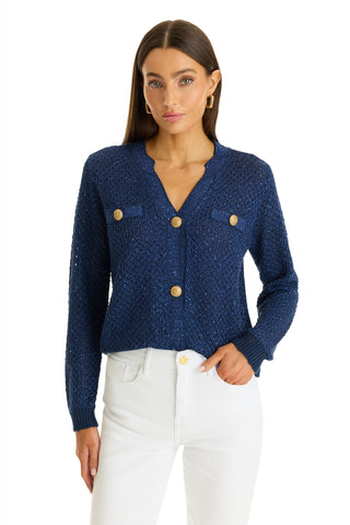A woman with straight brown hair wears the Shannon Sequin Cardigan Top in navy, featuring gold dome buttons and front pockets, paired with high-waisted white pants. She stands facing forward with a neutral expression.