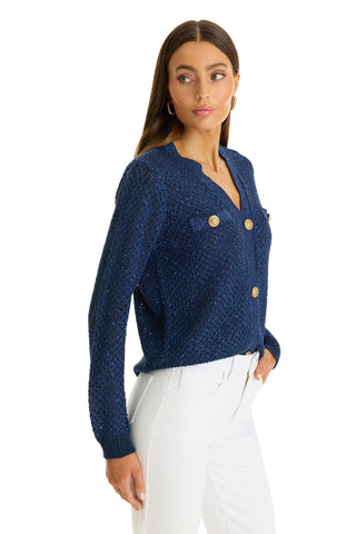 A woman with long brown hair wears the Shannon Sequin Cardigan Top in navy, featuring gold dome buttons and pockets. She pairs it with high-waisted white pants and stands sideways against a white background, looking slightly to the left.