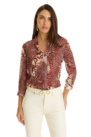 A woman wears the Sara Chiffon Blouse - Garnet Crush, featuring a maroon and white pattern with a high-low hem, tucked into high-waisted cream pants with a belt, standing against a plain white background.