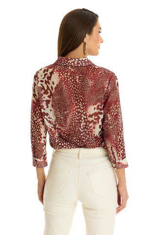 A woman with long brown hair, seen from the back, wears the Sara Chiffon Blouse in Garnet Crush with rolled sleeves and high-waisted white pants. She accessorizes with gold hoop earrings and looks to her left.