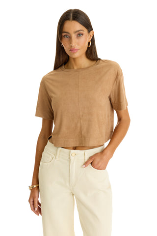 A woman with long brown hair wears the Naya Suede T-Shirt in Cafe, styled cropped, and high-waisted off-white pants. She stands with one hand in her pocket against a white background.
