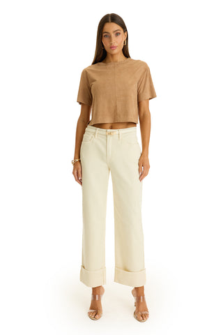 A woman faces forward in the Naya Suede T-Shirt - Cafe, paired with high-waisted cream wide-leg cuffed pants and clear-heeled sandals. She has straight hair, minimal accessories, and stands against a white background.