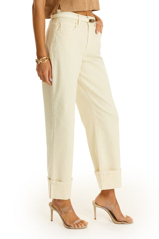 Wearing the Ren Cropped Cuff Jean in Ivory, a woman pairs them with a tan crop top, gold bracelet, and beige high-heeled sandals, posing with one hand in her pocket against a white background.