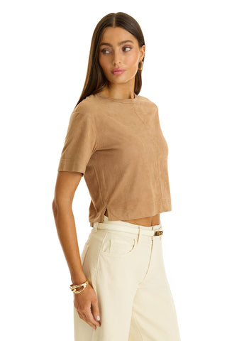 A woman with long brown hair wears the Naya Suede T-Shirt - Cafe, paired with high-waisted off-white pants and gold jewelry, standing against a white background and looking stylishly to her left.
