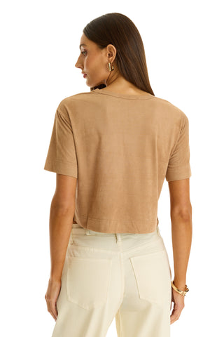 A woman with long brown hair stands facing away, wearing the Naya Suede T-Shirt - Cafe in a tan cropped fit and high-waisted off-white jeans. She accessorizes with hoop earrings and a bracelet—a perfect layering piece.
