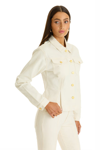 A woman with long brown hair wears the Bethanie Denim Jacket in ivory with gold buttons and matching pants. She stands sideways, one hand on her hip, looking off to the side against a white background.