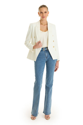 A person stands facing forward in the Sasha Double Breasted Blazer - White over a white top, blue jeans, beige heeled sandals, gold earrings, and a gold necklace against a plain white background.