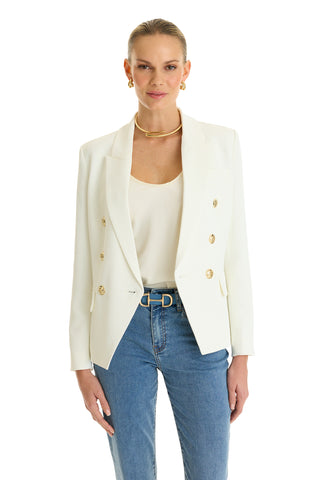 A woman stands smiling against a plain white background, wearing the Sasha Double Breasted Blazer in white over a cream top and blue jeans, styled with gold hoop earrings and a gold necklace.