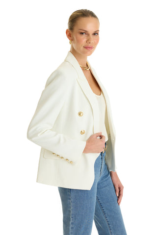 A woman, smiling with her hair pulled back, wears the Sasha Double Breasted Blazer in white paired with a white top and blue jeans as she stands against a white background.