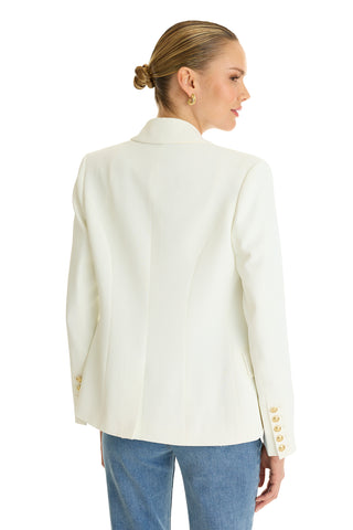 A woman with slicked-back hair models the Sasha Double Breasted Blazer in white with gold sleeve buttons and blue jeans. She is turned away, looking to the side, highlighting the back of the blazer.