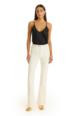 A woman stands against a white background wearing a black Yara Silk Camisole with a deep v-neck, high-waisted cream pants with gold buttons at the hem, gold open-toe heels, and hoop earrings.