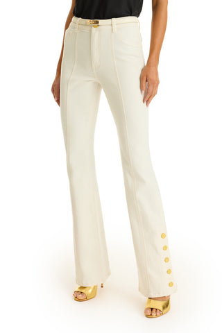 Someone wears the Veronica Button Jean in ivory, featuring gold buttons near one leg's hem, paired with a black top and gold open-toe high heels.