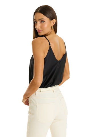 A woman with straight brown hair wears the Yara Silk Camisole - Black, paired with off-white pants, posing with her back slightly turned and glancing over her shoulder against a white background.