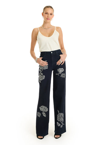A woman poses against a white background wearing the Kate Crystal Rose Jean in indigo, featuring high-waisted, wide-leg denim with white floral embroidery. She pairs them with a white sleeveless top and black open-toe heels, hands in her pockets.