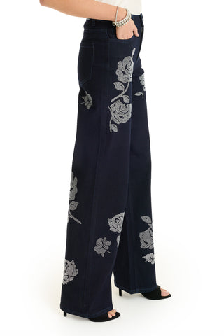 Someone wearing the Kate Crystal Rose Jean - Indigo, featuring wide legs and sparkling floral details, paired with black heeled sandals and a silver bracelet. Only their lower half is visible against a white background.