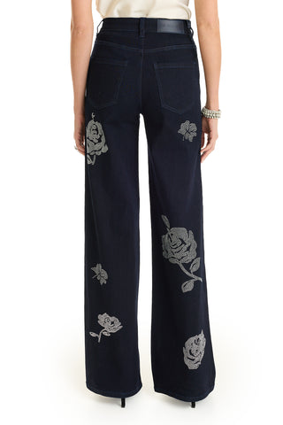 A person stands with their back to the camera wearing the Kate Crystal Rose Jean - Indigo, featuring silver rose embroidery on the back pockets, legs, and lower hem.