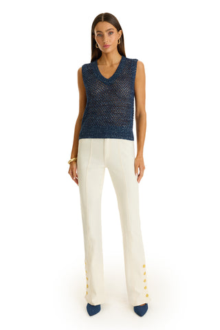 A woman stands against a white background wearing the Addison Sequin Crochet Top in navy, a sleeveless knit paired with white high-waisted pants with gold hem buttons, blue pointed-toe shoes, and gold jewelry.