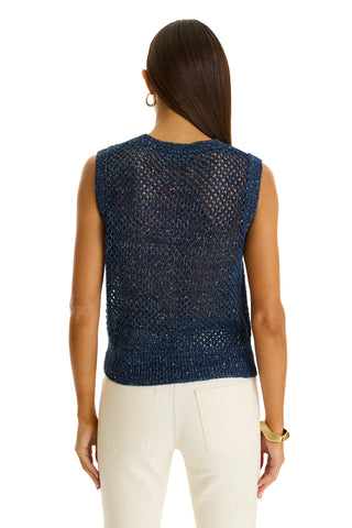 A woman with straight brown hair, wearing a sleeveless navy Addison Sequin Crochet Top and white pants, stands with her back to the camera, accessorized with a gold bracelet and hoop earrings for a resort-ready look.