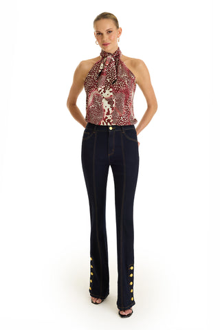 A woman poses against a white background wearing the Rina Tie Neck Blouse in Garnet Crush, a sleeveless halter-neck top with a red and white pattern, paired with high-waisted dark blue jeans featuring yellow buttons on the lower legs.