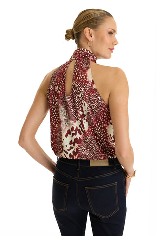 A woman with blonde hair in a low bun wears the Rina Tie Neck Blouse - Garnet Crush, featuring a maroon and white abstract print, sleeveless design, and open back detail. She pairs it with dark blue jeans and stands turned slightly to the side.