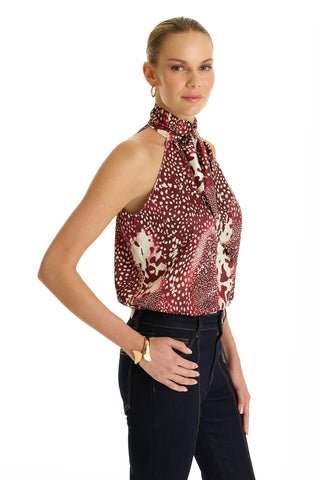 A woman poses against a white background, wearing the Rina Tie Neck Blouse in Garnet Crush paired with dark jeans, a gold bracelet, and hoop earrings. Her hair is tied back and she has a slight smile.