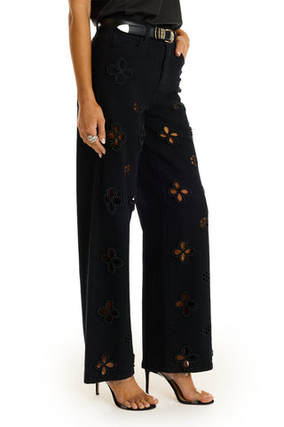 A person wearing Kaia Pearl High Rise Jean - Black with floral cutout designs revealing skin, paired with a black belt featuring a gold buckle and black high-heeled sandals.