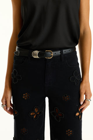 A person wearing a sleeveless black top, black jeans with cut-out flower patterns and studs, accessorized with the Blair Belt - Black featuring mixed metal details, and sporting multiple rings.