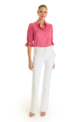 A woman wears the Cindy Ruffle Sleeve Shirt in Petal Pink with puffed sleeves, high-waisted white pants with gold buttons, open-toed heels, her hair tied back, and accessorizes with hoop earrings and a necklace.