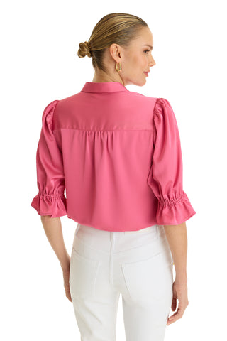 A woman with blonde hair in a low bun wears the Cindy Ruffle Sleeve Shirt in Petal Pink with puffed sleeves and white pants, standing with her back to the camera.