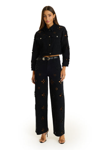 A woman with long brown hair and a neutral expression faces forward, wearing the Kaia Pearl Jean Jacket in black, wide-leg floral cutout pants, a black belt with a gold buckle, and black open-toe heels.