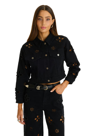 A woman with long brown hair models the Kaia Pearl Jean Jacket in black, featuring embellished details and posing with one hand in her pocket against a white background.