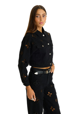 A woman with long brown hair wears the Kaia Pearl Jean Jacket in black and matching black pants with floral cutouts. She stands against a white background, gazing at the camera with one hand in her pocket.