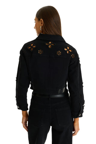 A person with straight brown hair stands facing away from the camera, wearing black pants and the Kaia Pearl Jean Jacket - Black, featuring floral cutout designs and embellishments on the back and sleeves.