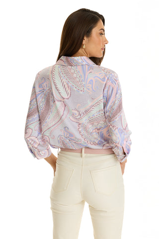 A woman with long brown hair models the Sara Silk Blouse - Pastel Paisley, paired with high-waisted white pants, standing with her back to the camera and head turned to the side against a white background.