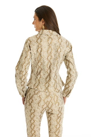 A woman with long brown hair wears gold hoop earrings and the Bethanie Denim Jacket in Gold Python, paired with matching pants, standing against a white background.