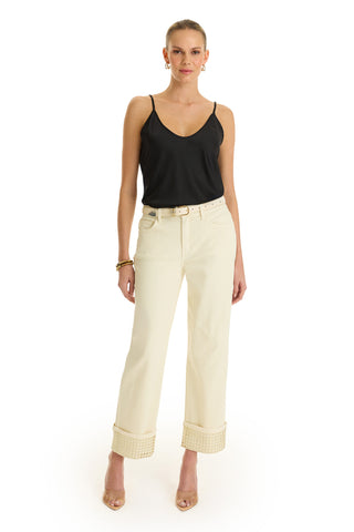 A woman faces forward in a black sleeveless top, gold hoop earrings, bracelets, nude heels, and the Ren Cropped Cuff Jean - Gold Mirror—cream-colored cropped jeans with four-way stretch denim. The background is plain white.