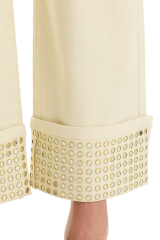 Close-up of the Ren Cropped Cuff Jean - Gold Mirror, featuring cream-colored denim with wide, cropped cuffs adorned with rows of gold metallic grommets. A portion of the wearer’s ankle and foot is visible below the hem.