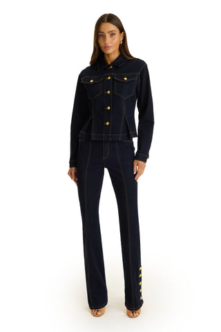 A woman with long brown hair faces forward against a white background, wearing the Bethanie Denim Jacket - Indigo with a subtle peplum hem and gold buttons, paired with matching dark denim pants and nude heels.