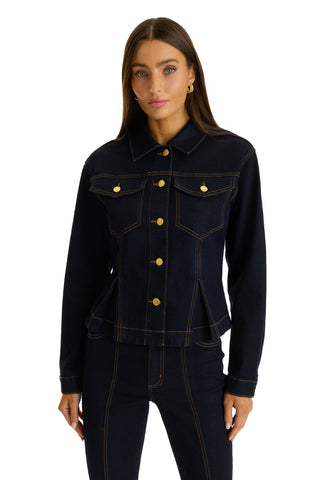 A woman wearing the Bethanie Denim Jacket - Indigo, a fitted dark denim jacket with gold buttons, and matching dark denim pants stands against a white background.