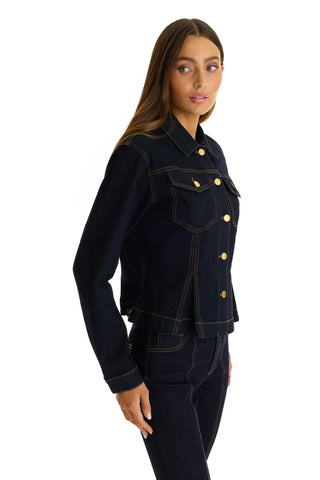 A woman with long brown hair models the Bethanie Denim Jacket in Indigo, featuring gold buttons and paired with matching jeans. She stands sideways, looking over her shoulder against a plain white background.