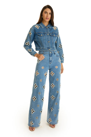 A woman with long brown hair poses with one hand in her pocket, wearing the Kaia Pearl High Rise Jean in medium blue—a pair of straight-leg jeans featuring bold white and beige floral appliqués.