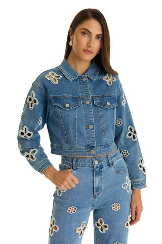 A woman wears the Kaia Pearl Jean Jacket in medium blue, styled with matching high-waisted jeans featuring white and brown floral cutout designs. She has long brown hair, neutral makeup, and stands confidently with one hand on her hip.