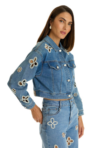 A woman poses with one hand on her hip against a white background, wearing the Kaia Pearl Jean Jacket in medium blue and matching jeans, both featuring white lace flower cut-out embellishments.