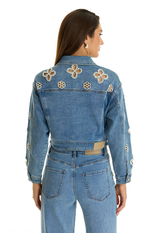 A woman with straight brown hair wears the Kaia Pearl Jean Jacket in medium blue, featuring embellished details, paired with matching high-waisted jeans against a white background.