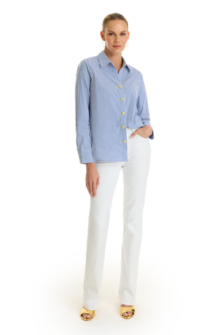 A woman stands against a white background, wearing the Jasmine Shirt - Blue Stripe paired with white pants, gold hoop earrings, and yellow open-toe heels. Her hair is pulled back and one hand rests in her pocket.