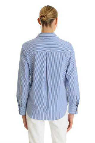 A person with blonde hair in a bun is shown from behind wearing the Jasmine Shirt - Blue Stripe, a long-sleeve, blue-and-white striped stretch poplin shirt, paired with white pants against a white background.