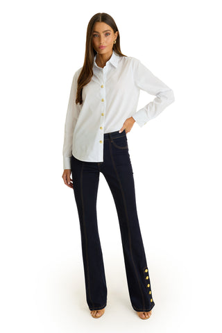A woman faces forward, wearing the Jasmine Shirt - White with a pleated back yoke and dark flared jeans featuring gold buttons. She has long brown hair, neutral heels, and poses against a plain white background.