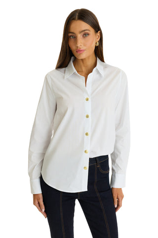 A woman with long brown hair wears the Jasmine Shirt - White, featuring a pleated back yoke, paired with dark jeans. She stands against a white background with her left hand in her pocket.